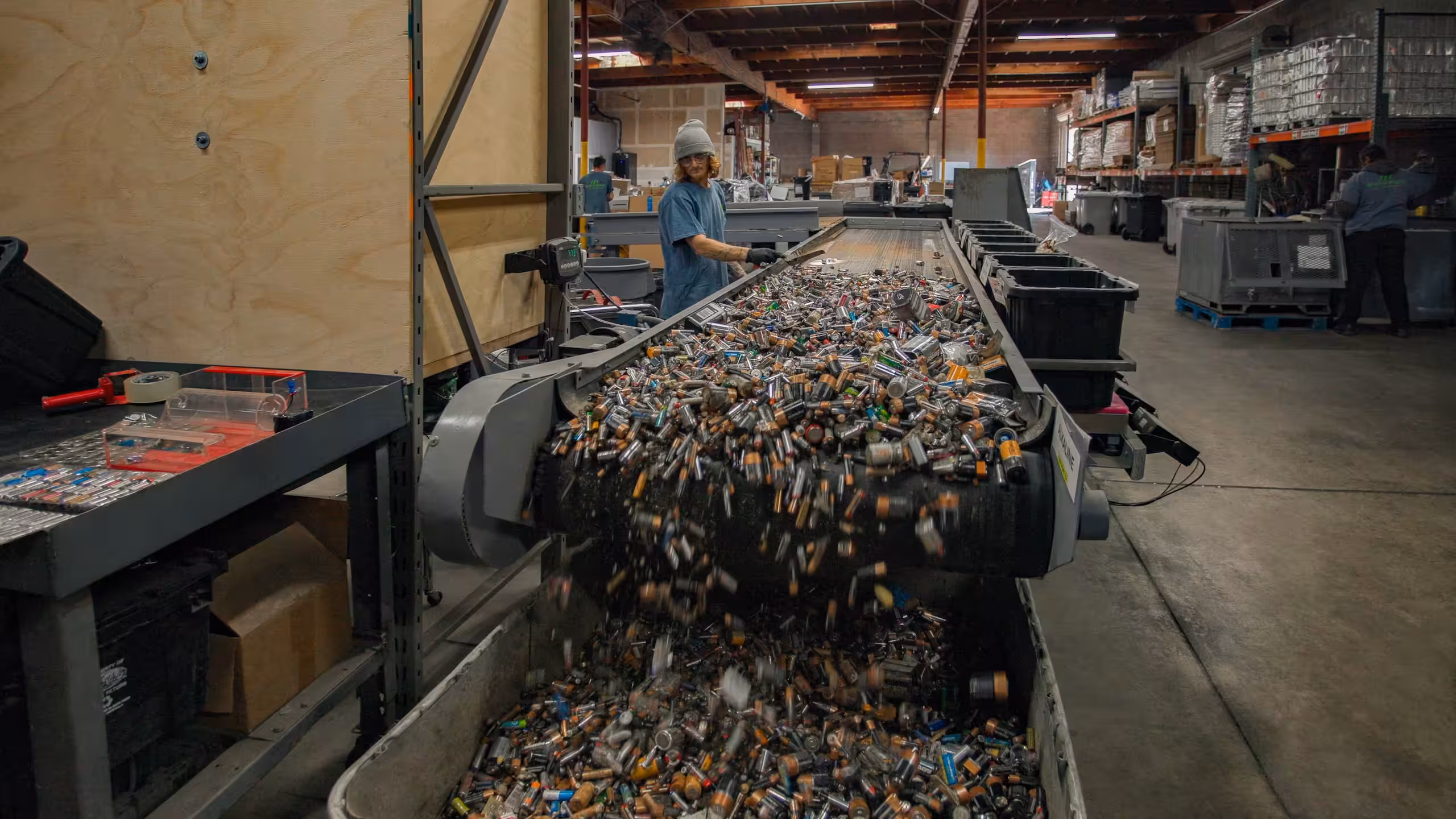 Worker sorting a large conveyor belt full of batteries inside a recycling facility warehouse.