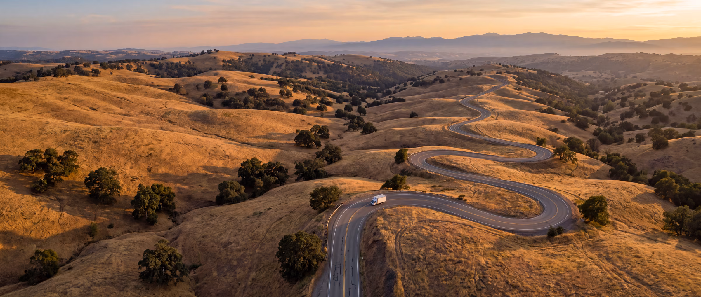 A white box truck on a road winding through golden California foothills at dusk